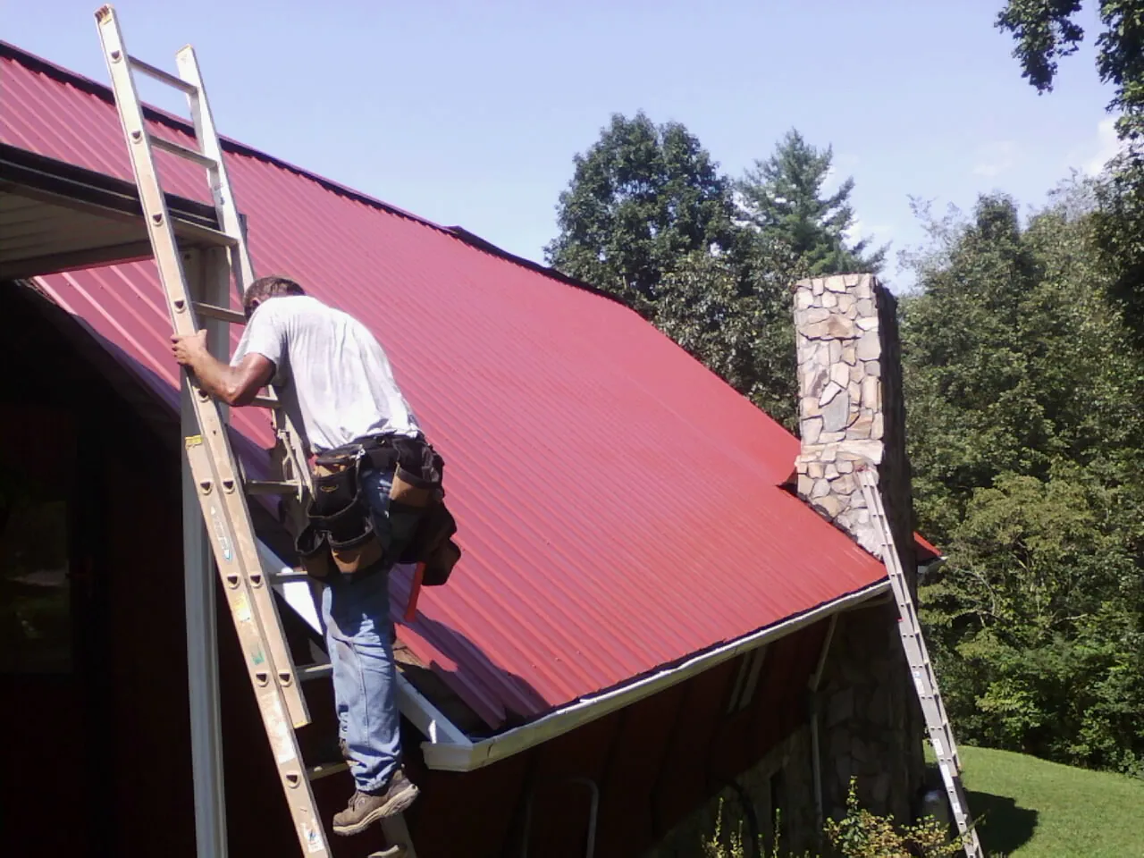 Roofer on a ladder beside a red metal roof and stone chimney — Soffit & Fascia Repair by Roofing Brothers in Marshville